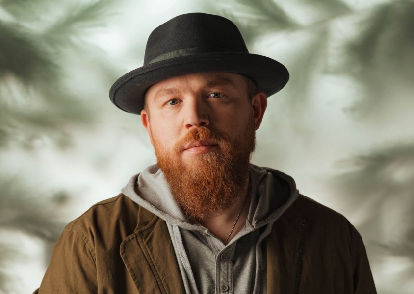A man with a ginger beard wearing a black hat, grey jumper and green jacketx